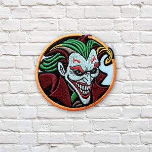 May include: Circular embroidered patch of the Joker character. The patch has a black border with an orange outline. The Joker's face is shown with green hair, white skin, and a red suit. The patch is set against a white brick wall.