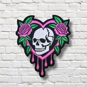 May include: Embroidered patch featuring a white skull within a pink heart shape, adorned with pink roses and green leaves. The heart has pink dripping accents. The patch is set against a white brick wall.