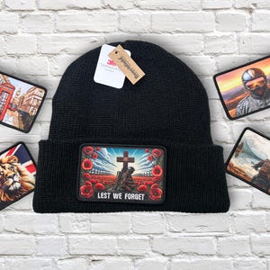 May include: Black knitted beanie hat with a rectangular patch depicting a soldier at a cross in a field of red poppies, with the text "Lest We Forget." Patches with images of London, a lion, and a motorcyclist surround the hat.