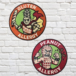 Puede incluir: Dos parches redondos bordados con personajes de dibujos animados. Uno dice "ALERGIA AL GLUTEN" y presenta un personaje con cabeza de trigo sosteniendo una baguette. El otro dice "ALERGIA AL CACAHUETE" y muestra un personaje con forma de cacahuete.
