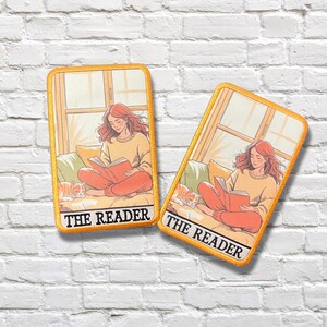May include: Two rectangular patches with a yellow border. Each patch features an illustration of a person reading a book, with a cat nearby. The text "THE READER" is at the bottom. The background is a window and a couch.