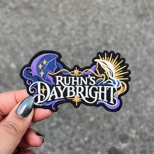 Puede incluir: Un parche bordado con fondo negro y el texto blanco RUHN'S DAYBRIGHT. El diseño incluye una silueta de perfil morada a la izquierda con estrellas amarillas y una silueta de perfil blanca a la derecha con rayos de sol amarillos.