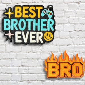 Puede incluir: Dos parches bordados sobre una pared de ladrillos blancos. El parche superior dice "BEST BROTHER EVER" con un icono de mando de juego y una carita sonriente. El parche inferior dice "BRO" en llamas.