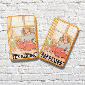 May include: Two embroidered patches featuring a woman reading a book, with a cat nearby. The patches have an orange border and the text "THE READER" at the bottom. The woman is wearing a gray sweater and orange pants.