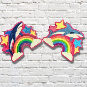 Puede incluir: Dos parches bordados con tiburones saltando sobre arcoíris coloridos. El parche izquierdo muestra un tiburón azul oscuro, mientras que el derecho muestra un tiburón azul claro. Ambos diseños incluyen estrellas amarillas, azules y nubes blancas.