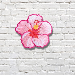 May include: An embroidered patch in the shape of a hibiscus flower. The flower features five petals with a gradient of white and light pink, accented by a bright red centre and a yellow stamen, all outlined with a vibrant hot pink thread border.