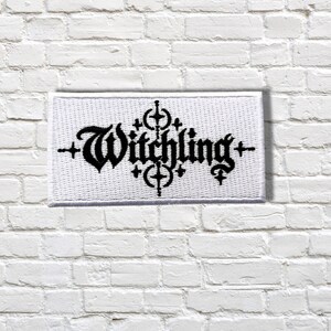 May include: A rectangular white embroidered patch featuring the black text Witchling in a gothic-style font. The design includes symmetrical black cross-like symbols positioned above and below the centre of the word, with smaller crosses on each side.