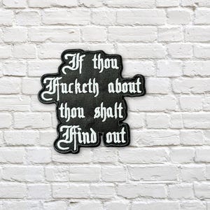 Peut inclure: Écusson noir brodé avec du texte blanc : « If thou fucketh about thou shalt find out ». L'écusson a une forme unique et est posé sur un mur de briques blanches.