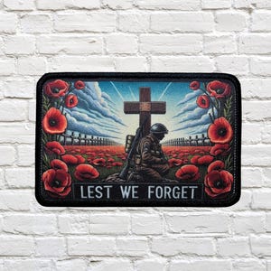 Op de afbeelding: Een zwart-witte patch met een knielende soldaat naast een houten kruis. De soldaat draagt een militair uniform en een helm. De achtergrond is een veld met rode klaprozen met de tekst "Lest We Forget".