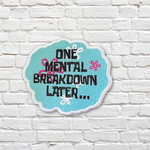 Pode incluir: Um patch redondo com o texto "ONE MENTAL BREAKDOWN LATER..." em preto, sobre um fundo azul claro com uma borda branca. O patch tem detalhes florais rosa e brancos e é colocado contra uma parede de tijolos brancos.