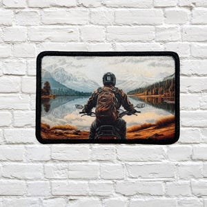Può includere: Toppa rettangolare raffigurante un motociclista di fronte a un lago e a un paesaggio montano. Il motociclista, visto da dietro, indossa un casco e una giacca. Il paesaggio, con alberi e montagne, si riflette nell'acqua. La toppa ha un bordo nero.
