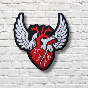 May include: Embroidered patch featuring a red anatomical heart with white wings, set against a black background. The patch has a black border and is designed to be sewn or ironed onto fabric. The wings are detailed with white stitching.
