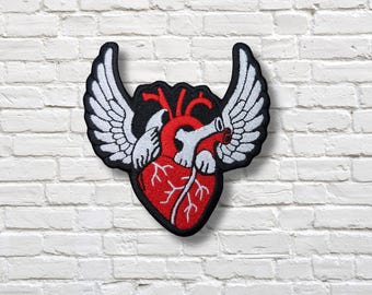 Op de afbeelding: Geborduurde patch met een rood anatomisch hart met witte vleugels, tegen een zwarte achtergrond. De patch heeft een zwarte rand en is ontworpen om op stof te worden genaaid of gestreken. De vleugels zijn gedetailleerd met witte stiksels.