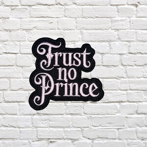 May include: A black and white embroidered patch with the text "Trust no Prince". The patch has a decorative border and is set against a white brick wall.
