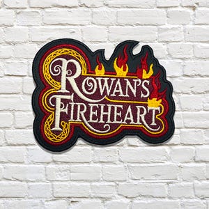 May include: Embroidered patch with the words "Rowan's Fireheart" in white and burgundy, surrounded by yellow and red flames. The patch has a black border and is set against a white brick wall.