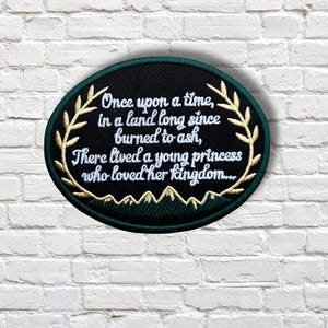 May include: Oval-shaped embroidered patch with the text "Once upon a time, in a land long since burned to ash, There lived a young princess who loved her kingdom..." in white script. The patch has a black background with gold accents.