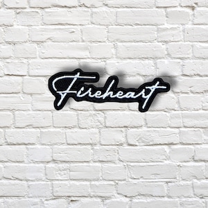 May include: A black patch with white embroidered text that reads "Fireheart" in a cursive font. The patch has a black border and is set against a white brick wall background.