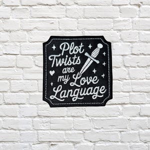 May include: Black patch with white text that reads "Plot Twists are my Love Language." A dagger graphic is in the center. The patch has a black border and is against a white brick wall.