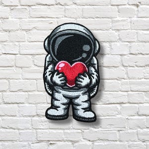 Puede incluir: Parche bordado con un astronauta de dibujos animados sosteniendo un corazón rojo. El astronauta lleva un traje espacial blanco con detalles en negro y gris. El parche está sobre una pared de ladrillos blancos.