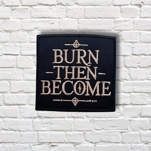 Puede incluir: Un parche cuadrado negro con el texto bordado "BURN -THEN- BECOME" en color beige claro. El parche tiene símbolos decorativos encima y debajo del texto. El parche está colocado sobre una pared de ladrillos blancos.
