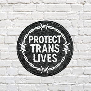 May include: A circular black patch featuring the white text "PROTECT TRANS LIVES" encircled by white barbed wire. The patch is set against a white brick wall.