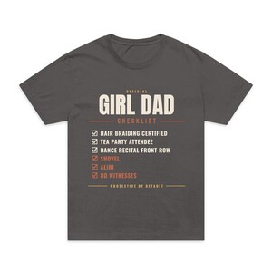 May include: A charcoal gray short-sleeve t-shirt featuring the text OFFICIAL GIRL DAD CHECKLIST in white and orange. The graphic includes a list of checked items: HAIR BRAIDING CERTIFIED, TEA PARTY ATTENDEE, DANCE RECITAL FRONT ROW, SHOVEL, ALIBI, NO WITNESSES.