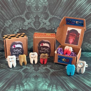 May include: A collection of tooth-shaped rings in various colors, including white, yellow, red, and blue, with some featuring rhinestones. The rings are displayed with small cardboard boxes, some with monkey images and the text "Curious Parcel."