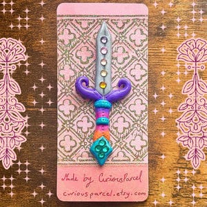 Könnte beinhalten: Eine dekorative schwertförmige Anstecknadel mit silberner Klinge und einem lila, orangefarbenen und türkisfarbenen Griff. Die Nadel befindet sich auf einer rosa Karte mit goldenem Muster und dem Text "Made by Curiousparcel curiousparcel.etsy.com".