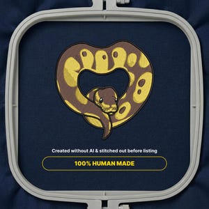 Könnte beinhalten: Ein Stickmotiv einer braun-gelben Schlange in Herzform, eingespannt in einen hellgrauen Stickrahmen auf dunkelblauem Stoff. Der Text darunter lautet: Created without AI & stitched out before listing 100% HUMAN MADE.