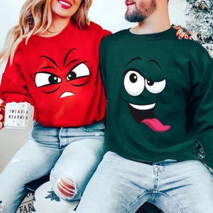 Könnte beinhalten: Zwei Sweatshirts, eines rot und eines grün, jeweils mit einem Cartoon-Gesicht. Das rote Sweatshirt hat ein wütendes Gesicht, während das grüne ein albernes Gesicht mit herausgestreckter Zunge hat. Die Person links hält einen Becher mit der Aufschrift "Sweater Weather".
