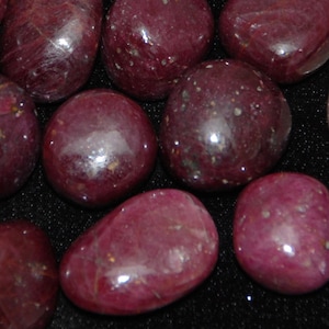 Könnte beinhalten: Eine Sammlung polierter, tiefroter Rubin-Edelsteine. Die Steine variieren in Größe und Form und haben eine glatte, reflektierende Oberfläche. Die Rubine haben eine satte, gesättigte Farbe mit subtilen Farbnuancen. Die Edelsteine sind auf einem dunklen Hintergrund verstreut.