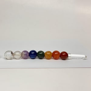 May include: A wand featuring a row of eight spherical beads in clear, purple, blue, green, yellow, orange, and red colors, capped with a clear crystal point. A silver-colored coin is placed to the left for scale, showing its circular face.