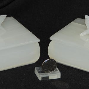 Op de afbeelding: Twee witte, boekvormige dozen met duivenbeeldjes. De dozen zijn gemaakt van een doorschijnend materiaal en hebben afgeronde randen. Tussen de dozen staat een kleine, heldere standaard met een munt.