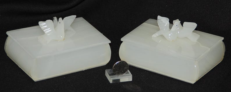 Op de afbeelding: Twee witte, boekvormige dozen met duivenbeeldjes. De dozen zijn gemaakt van een doorschijnend materiaal en hebben afgeronde randen. Tussen de dozen staat een kleine, heldere standaard met een munt.