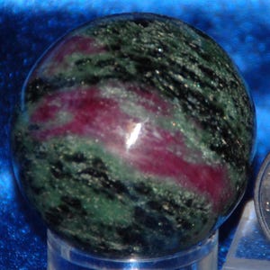 May include: A polished gemstone sphere with a mix of colors. The sphere has a base of green and black with streaks of deep red. The stone is smooth and round, and it sits on a clear stand. The overall effect is one of natural beauty.