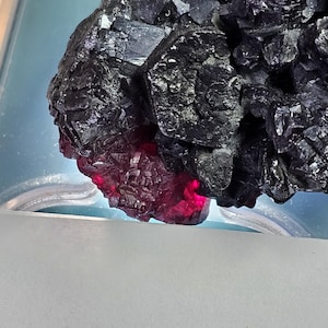 May include: Close-up of a dark, crystalline mineral formation with a deep black colour. The lower portion of the mineral glows with a vibrant red hue, creating a striking contrast. The mineral is set against a light blue background.