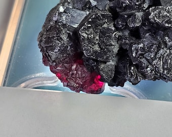 Könnte beinhalten: Nahaufnahme einer dunklen, kristallinen Mineralformation mit tiefschwarzer Farbe. Der untere Teil des Minerals leuchtet in einem leuchtenden Rotton und erzeugt einen auffälligen Kontrast. Das Mineral ist vor einem hellblauen Hintergrund platziert.