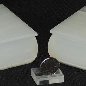 Op de afbeelding: Twee cr&egrave;mekleurige, boekvormige dozen met duifvormige dekselornamenten. Een munt rust op een kleine, transparante, rechthoekige standaard tussen de dozen. De dozen hebben afgeronde randen en een gladde, doorschijnende uitstraling.