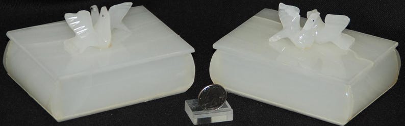 Op de afbeelding: Twee cr&egrave;mekleurige, boekvormige dozen met duifvormige dekselornamenten. Een munt rust op een kleine, transparante, rechthoekige standaard tussen de dozen. De dozen hebben afgeronde randen en een gladde, doorschijnende uitstraling.