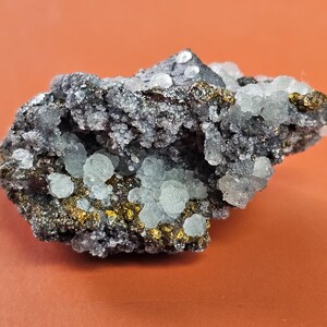Puede incluir: Primer plano de un espécimen mineral con cúmulos cristalinos azul claro sobre una matriz gris oscuro. El espécimen tiene parches dorados y se encuentra sobre un fondo naranja.
