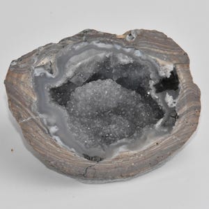 May include: A geode cut in half, revealing a cavity filled with sparkling, light gray crystals. The outer shell is a mix of brown, gray, and white bands. The overall shape is oval, with a rough, natural texture.