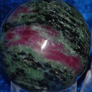 May include: A gemstone sphere with a polished surface, displaying a mix of deep green, black, and magenta hues in a swirling pattern. The sphere's smooth texture reflects light, highlighting the natural colours. A unique decorative item for the home.