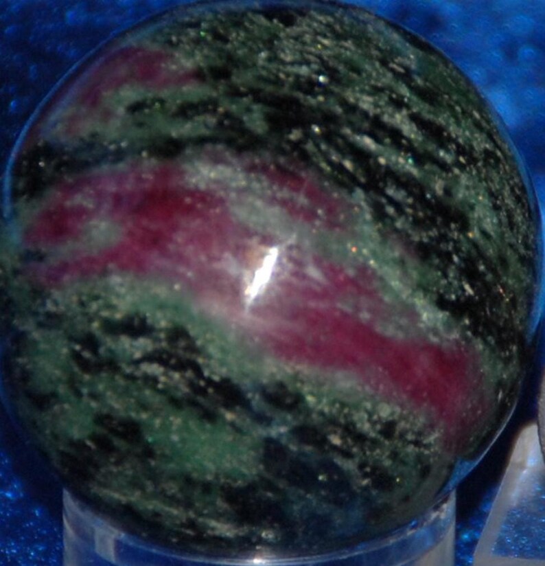 May include: A gemstone sphere with a polished surface, displaying a mix of deep green, black, and magenta hues in a swirling pattern. The sphere's smooth texture reflects light, highlighting the natural colours. A unique decorative item for the home.