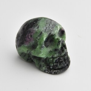 May include: A carved skull figurine made of green and black speckled stone. The skull has detailed features, including eye sockets, a nose cavity, and teeth. The stone has a polished finish.