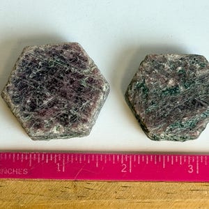 K&ouml;nnte beinhalten: Zwei hexagonale Rubinkristalle mit einer gesprenkelten dunkelroten und dunkelgr&uuml;nen Oberfl&auml;chenstruktur. Ein rosa Lineal liegt unter den Steinen und zeigt, dass der gr&ouml;&szlig;ere Kristall etwa 3,8 cm und der kleinere etwa 3,2 cm breit ist.