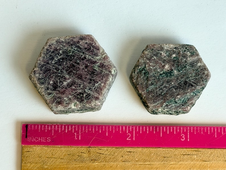 K&ouml;nnte beinhalten: Zwei hexagonale Rubinkristalle mit einer gesprenkelten dunkelroten und dunkelgr&uuml;nen Oberfl&auml;chenstruktur. Ein rosa Lineal liegt unter den Steinen und zeigt, dass der gr&ouml;&szlig;ere Kristall etwa 3,8 cm und der kleinere etwa 3,2 cm breit ist.