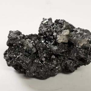 Puede incluir: Un espécimen mineral de color gris oscuro con formaciones cristalinas. La superficie rugosa y texturizada está salpicada de pequeños cristales brillantes y grupos de cristales translúcidos de color claro.
