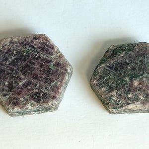 K&ouml;nnte beinhalten: Zwei rohe, sechseckige Mineralexemplare mit einer gesprenkelten Textur in tiefem Burgunderrot, Dunkelviolett und Waldgr&uuml;n. Die Oberfl&auml;chen dieser Steine sind rau und kristallin und weisen nat&uuml;rliche, geometrische Kanten auf.