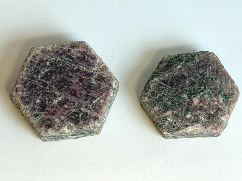 K&ouml;nnte beinhalten: Zwei rohe, sechseckige Mineralexemplare mit einer gesprenkelten Textur in tiefem Burgunderrot, Dunkelviolett und Waldgr&uuml;n. Die Oberfl&auml;chen dieser Steine sind rau und kristallin und weisen nat&uuml;rliche, geometrische Kanten auf.
