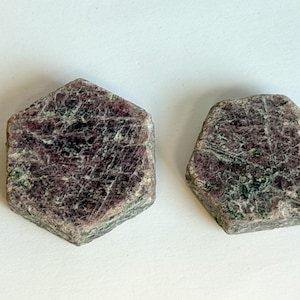 May include: Two raw, hexagonal-shaped ruby crystals with a deep maroon and dark red color palette. The surfaces of these stones exhibit a natural, textured appearance with visible mineral inclusions and a matte finish, showing distinct geometric edges.
